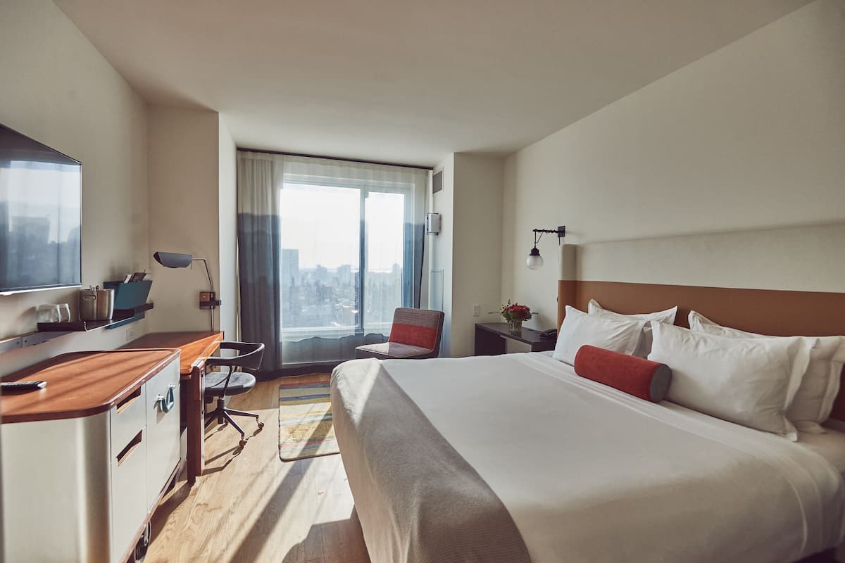 A hotel room with a large bed, a desk with a chair, a TV, and a view of the city through the window.
