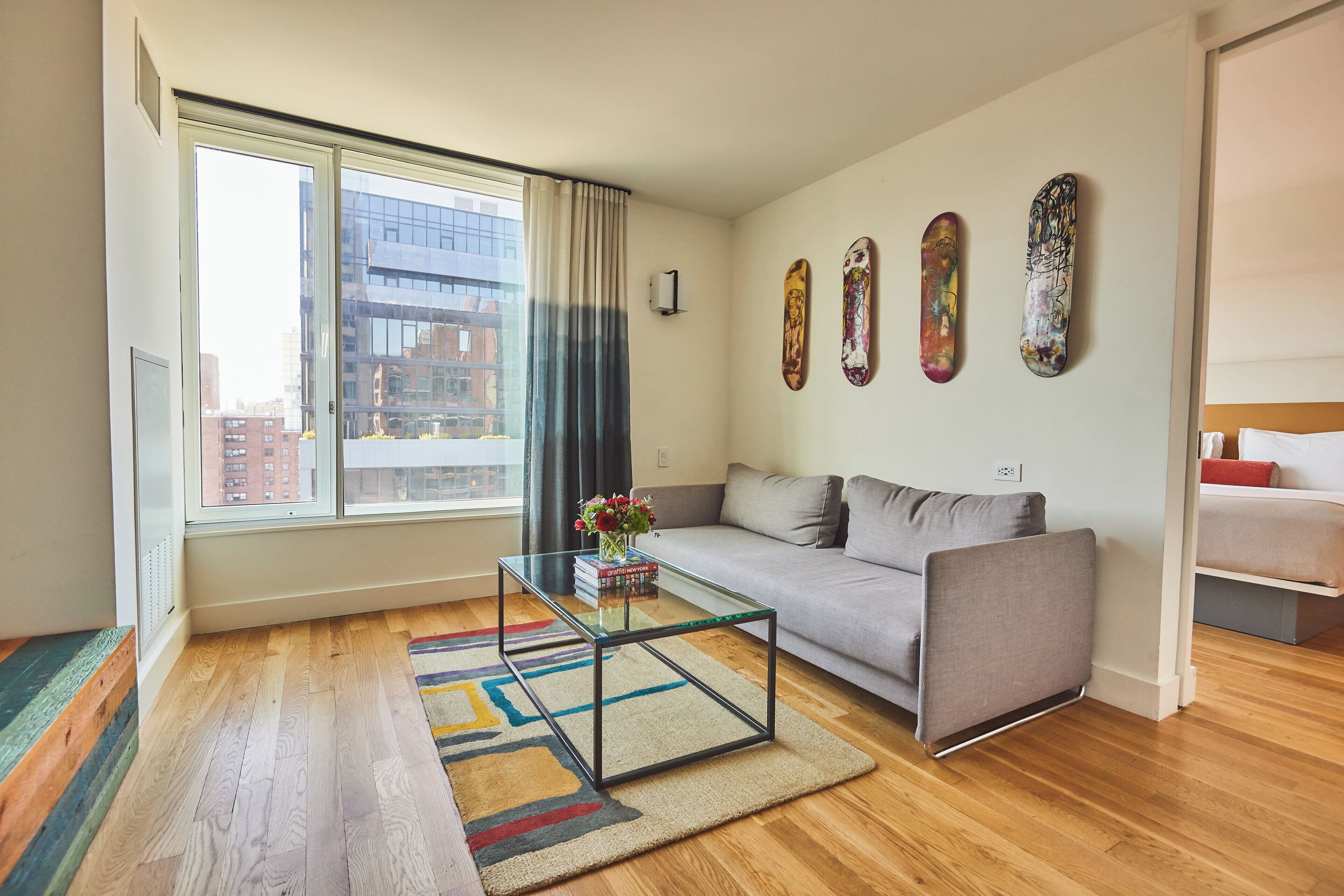 A modern living room with a glass coffee table, grey sofa, and a large window with curtains.