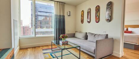 A modern living room with a glass coffee table, grey sofa, and a large window with curtains.