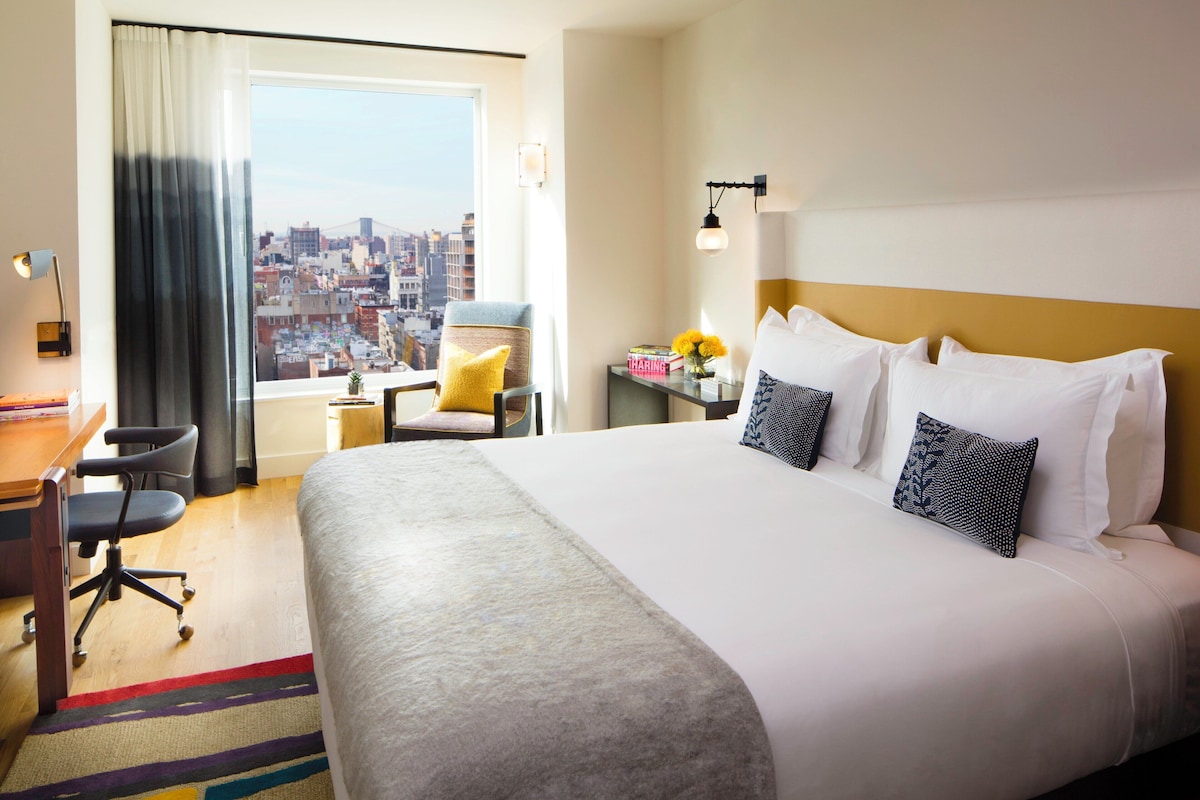 Habitación de hotel con una cama grande, un escritorio, una silla, una ventana con vista a la ciudad y una mesita de noche con lámpara.