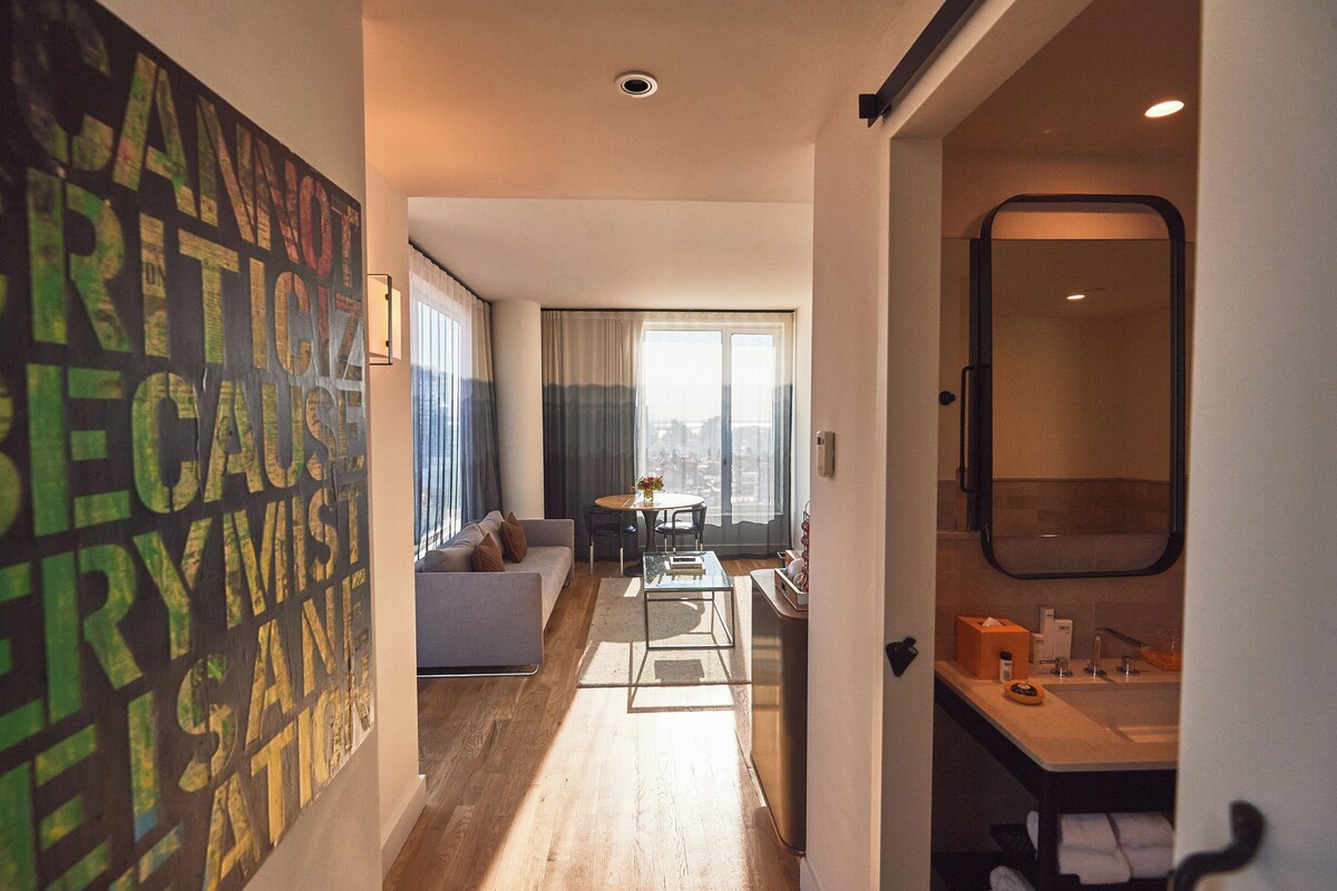 Habitación de hotel moderna con un ventanal grande, un sofá, una mesa de vidrio y un baño con espejo y lavamanos.