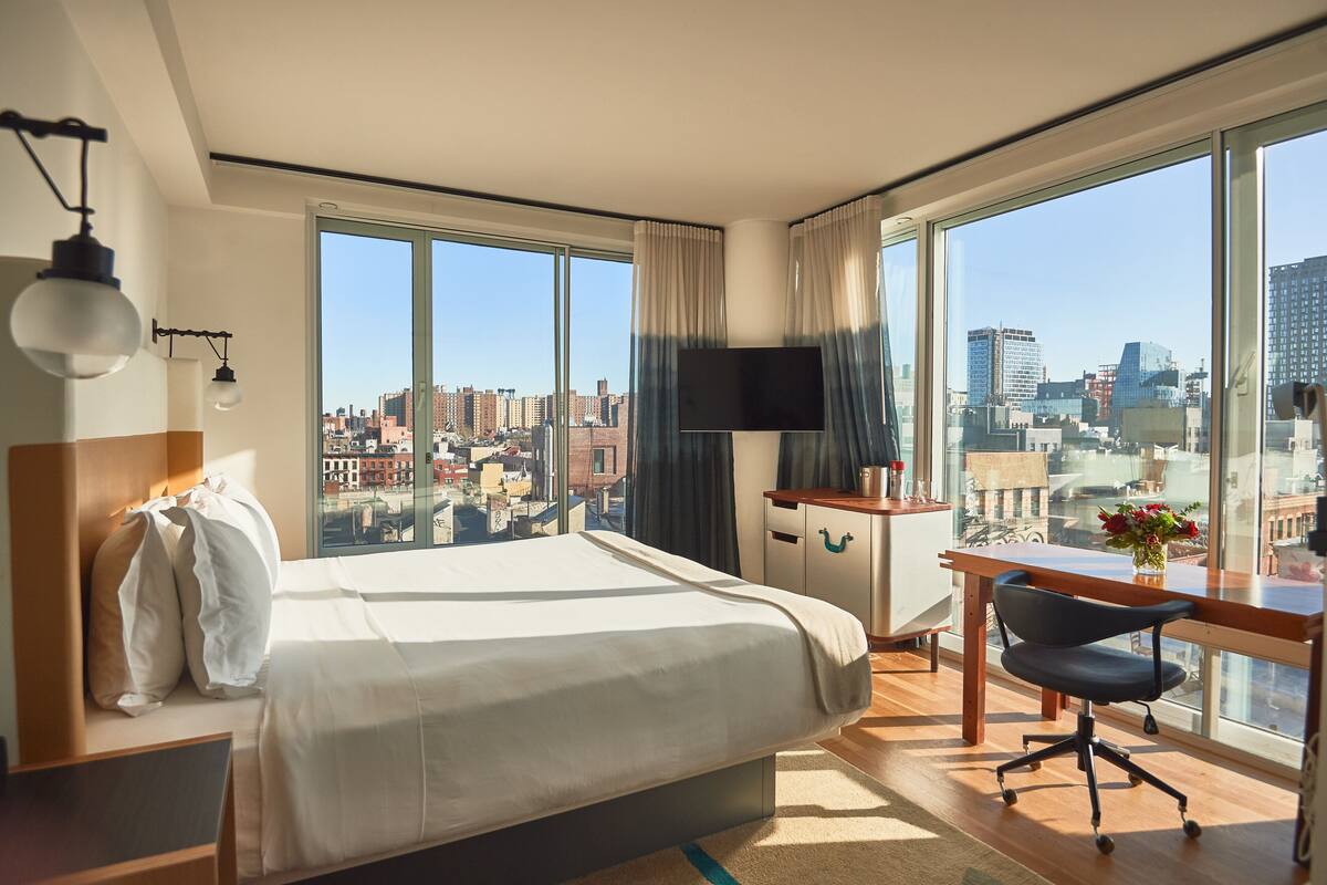 A hotel room with a large bed, a desk, a chair, and a TV. There is a view of the city through large windows.