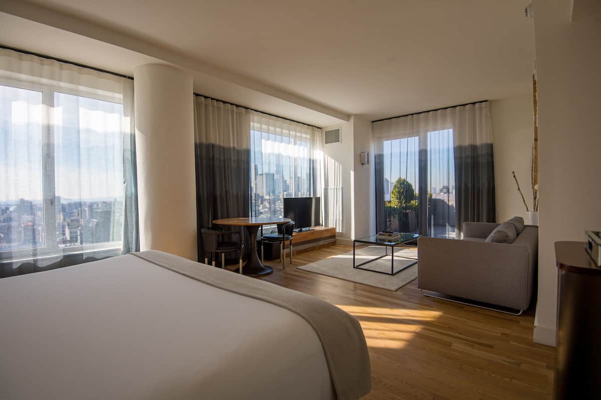 A modern hotel room with a large bed, a sofa, a dining table, and a TV. The room has large windows with curtains, offering a view of the city.