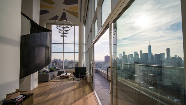 Een ruim balkon met uitzicht op de stadshemel, een modern woongedeelte met een bank en een eettafel, en een grote televisie aan de muur.