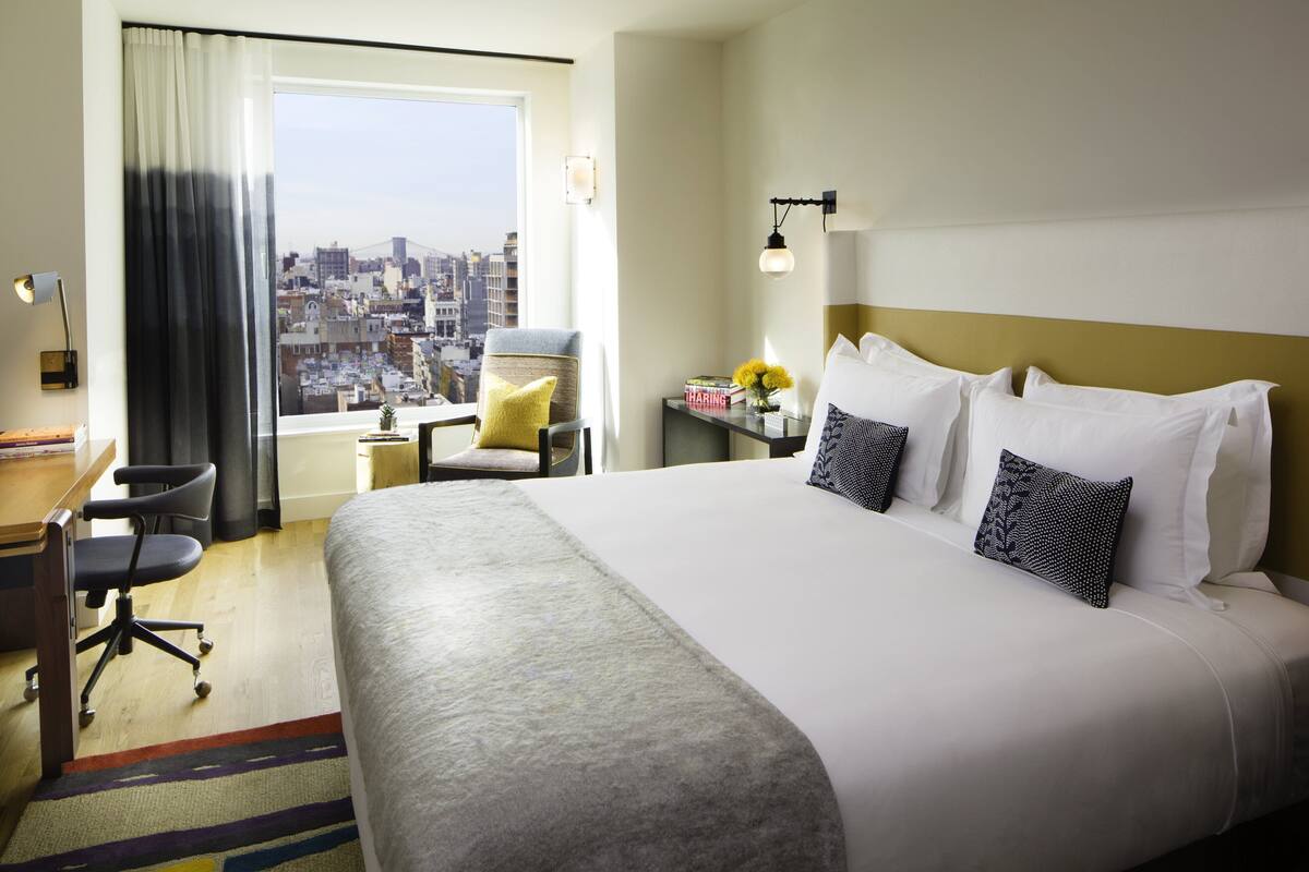 A hotel room with a large bed, a desk, a chair, a window with a city view, and a nightstand with a lamp.