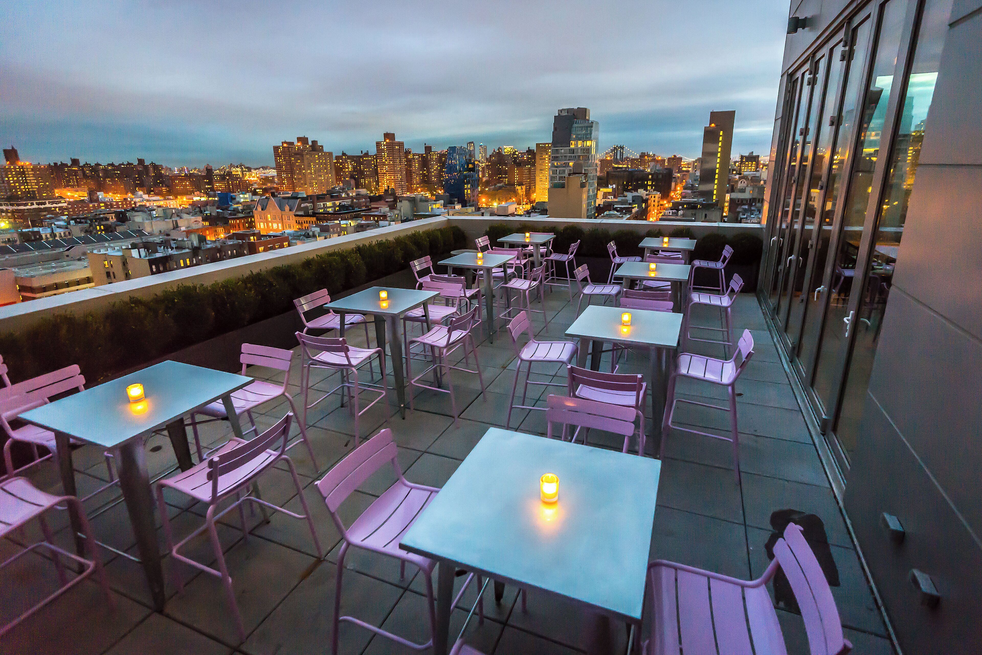 An outdoor terrace with tables and chairs, overlooking a cityscape at dusk.