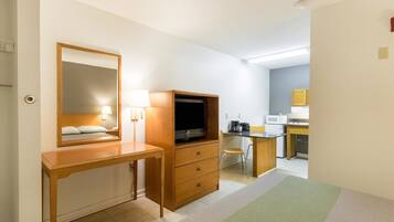 A hotel room with a wooden desk, a television, a mirror, a small kitchenette, and a green patterned rug.