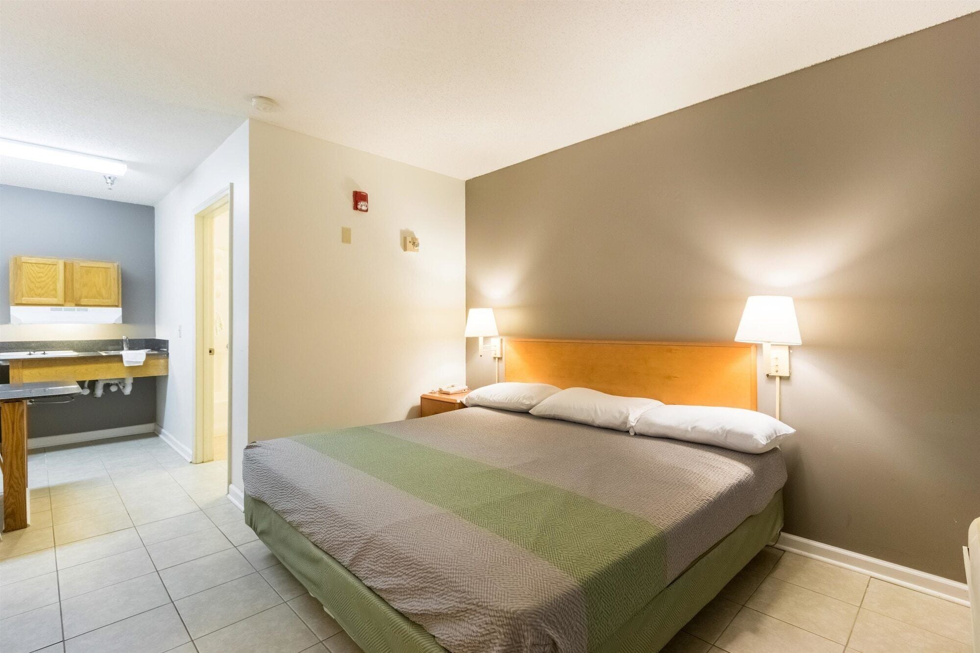 A hotel room with a bed, bedside lamps, a wooden headboard, a small bedside table, and a bathroom with a sink and mirror.