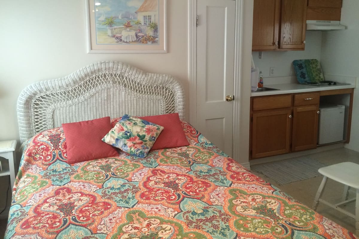 A bedroom with a patterned bedspread, a wicker headboard, a red pillow, and a painting of a seaside scene.