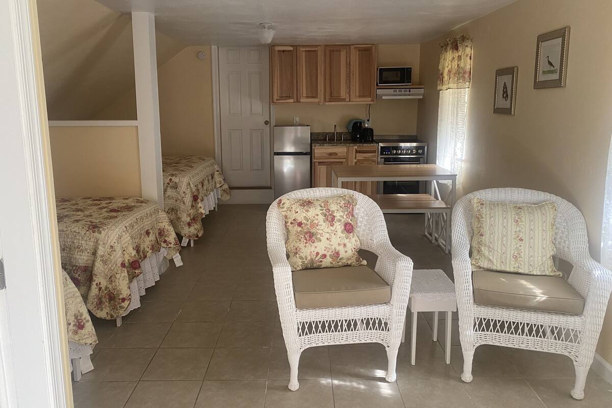 A compact living space with a kitchenette, two white wicker chairs with floral cushions, and a bed with floral bedding.