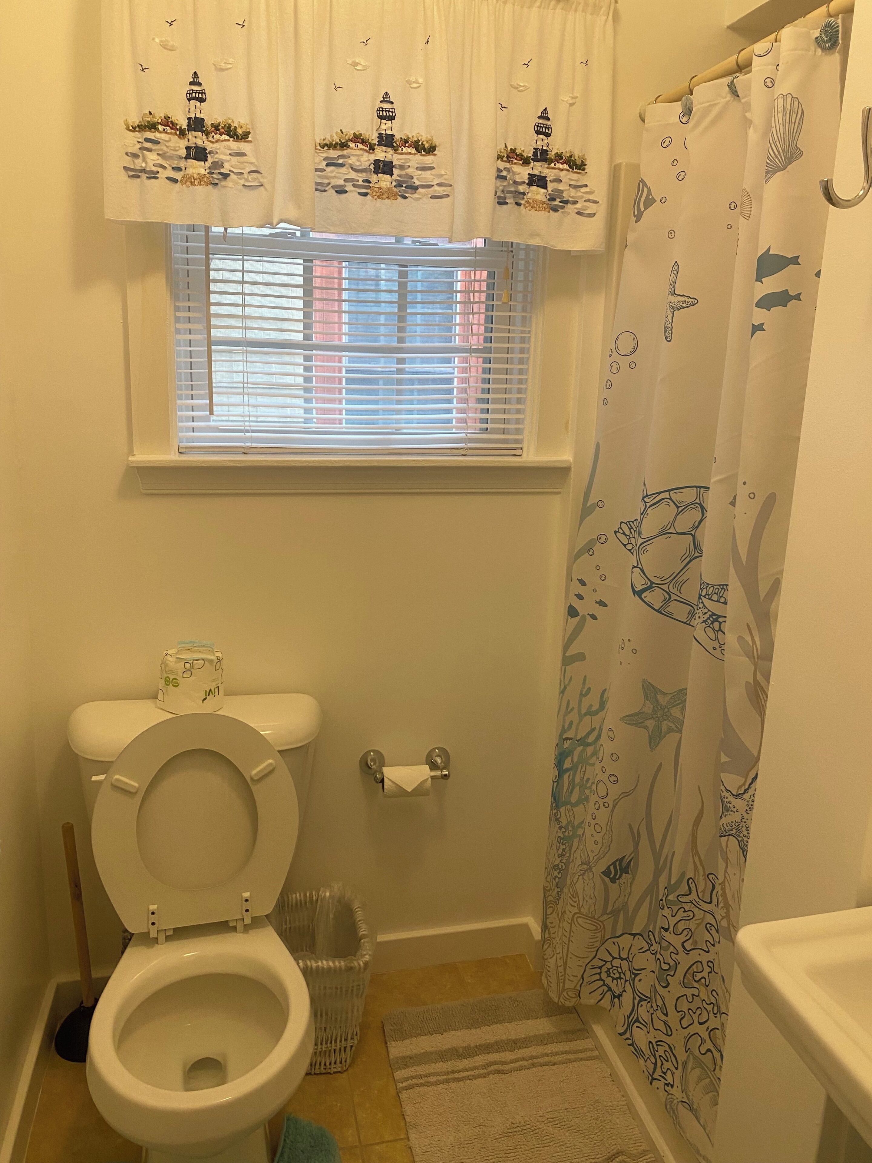 A bathroom with a toilet, a window with blinds, a shower curtain with marine life designs, and a waste basket.