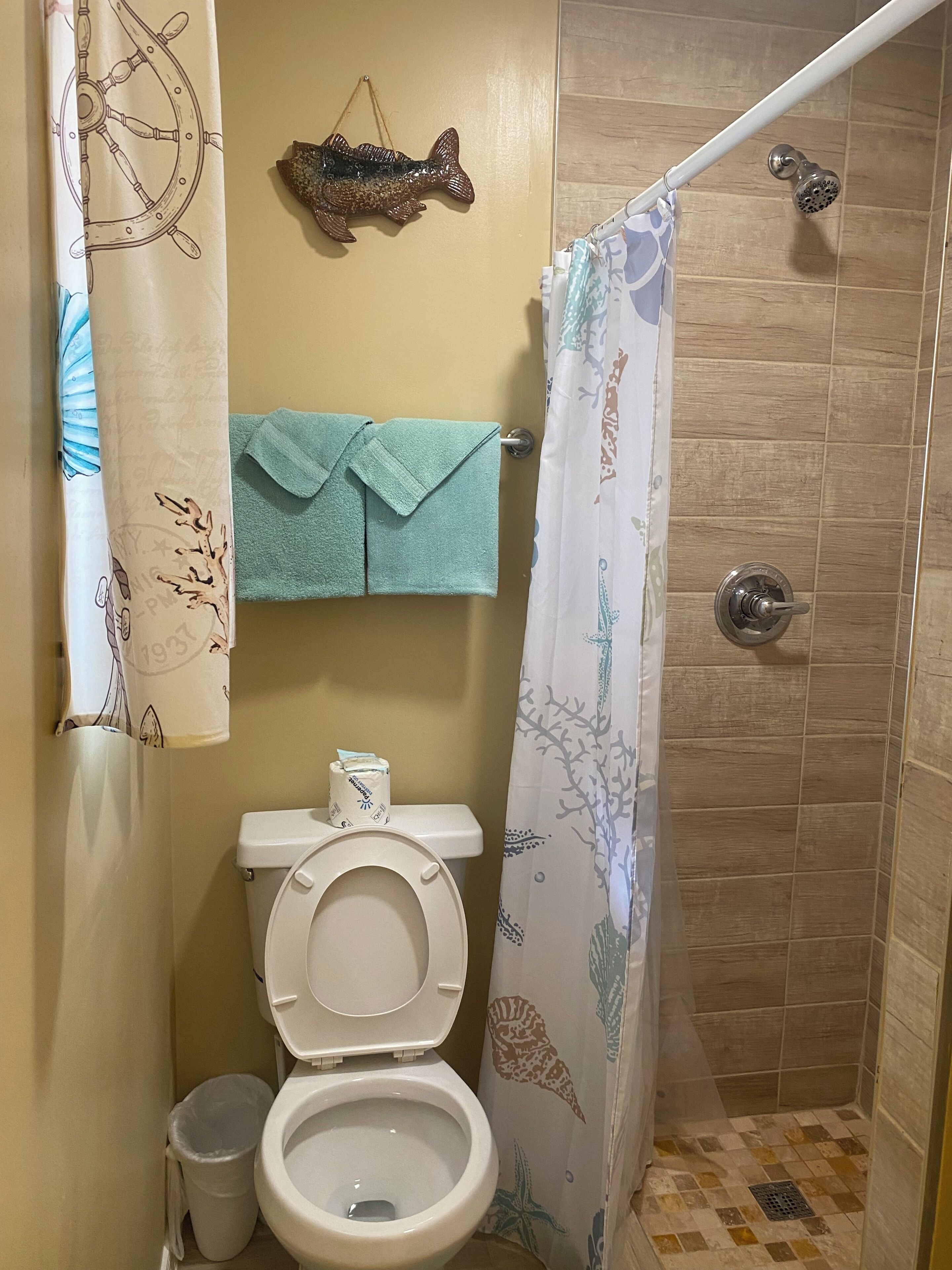 A bathroom with a toilet, a shower curtain with sea creature designs, a towel rack with two towels, and a decorative fish on the wall.