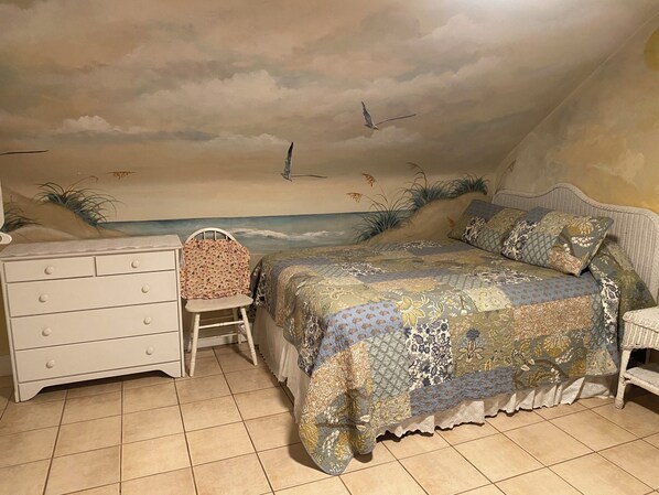 A bedroom with a bed, a dresser, a chair, and a wall mural of a beach scene. - Angie's Guest Cottage (Virginia Beach)