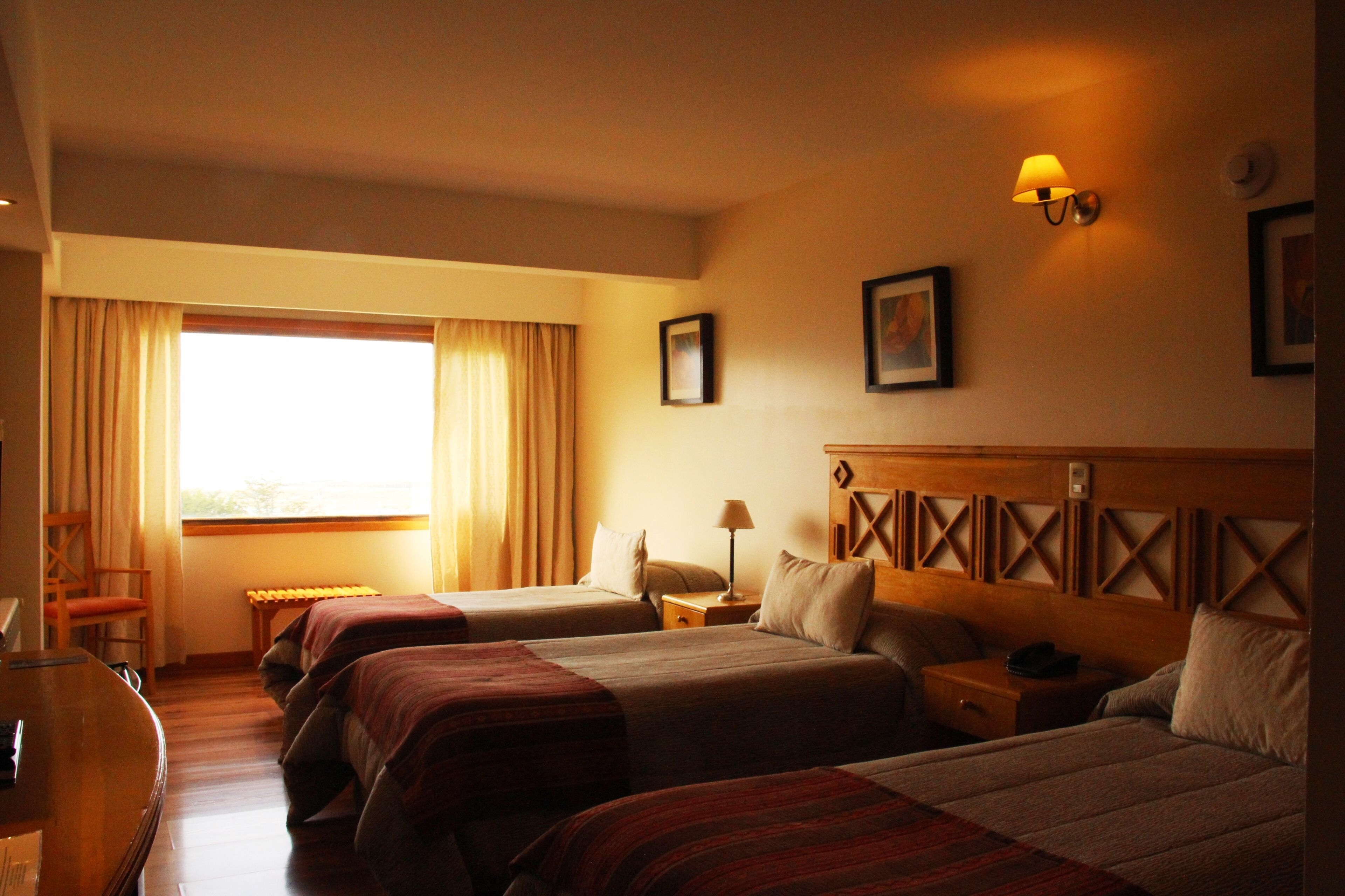 Quarto de hotel com três camas, uma mesa, uma cadeira e uma janela com cortinas.
