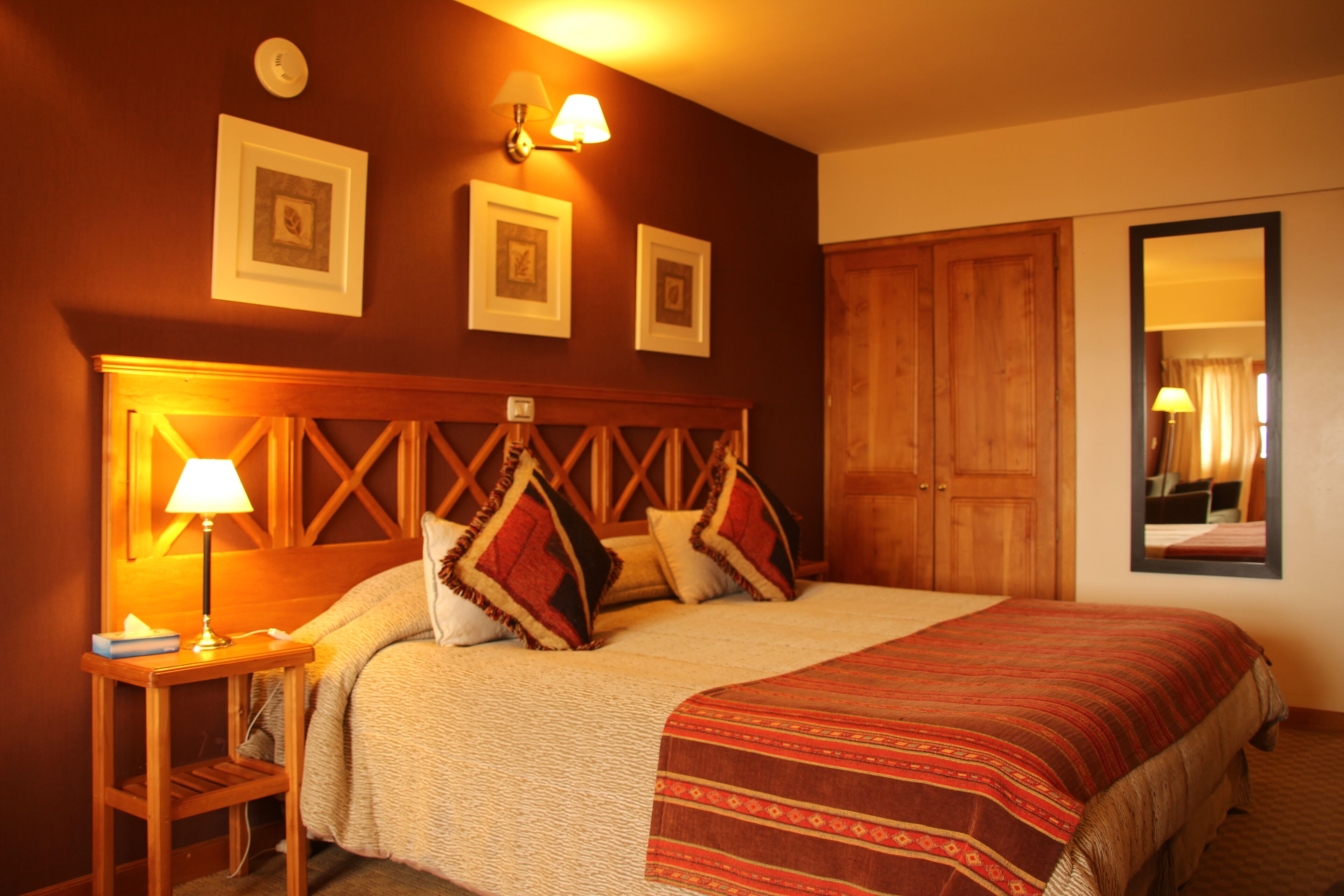 Quarto de hotel com cama, mesa de cabeceira com abajur, cabeceira de madeira e quadros nas paredes.