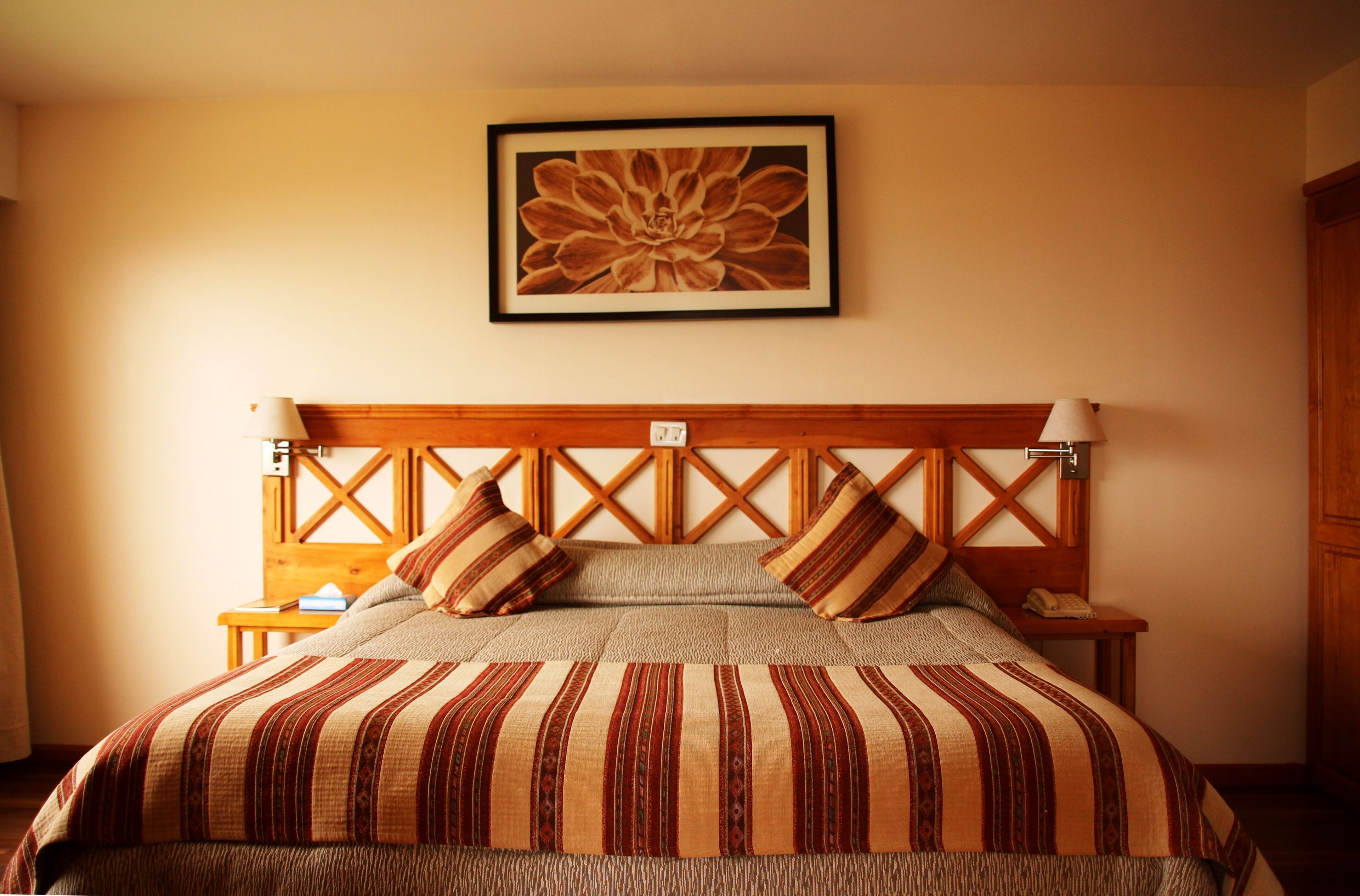 Quarto com cabeceira de madeira, cama com roupa de cama listrada, mesa de cabeceira com abajur e uma obra de arte floral emoldurada na parede.