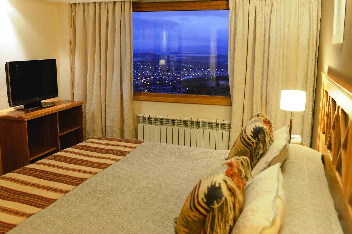 Une chambre d’hôtel avec un lit, un téléviseur posé sur un meuble, une fenêtre donnant sur la ville et un radiateur.
