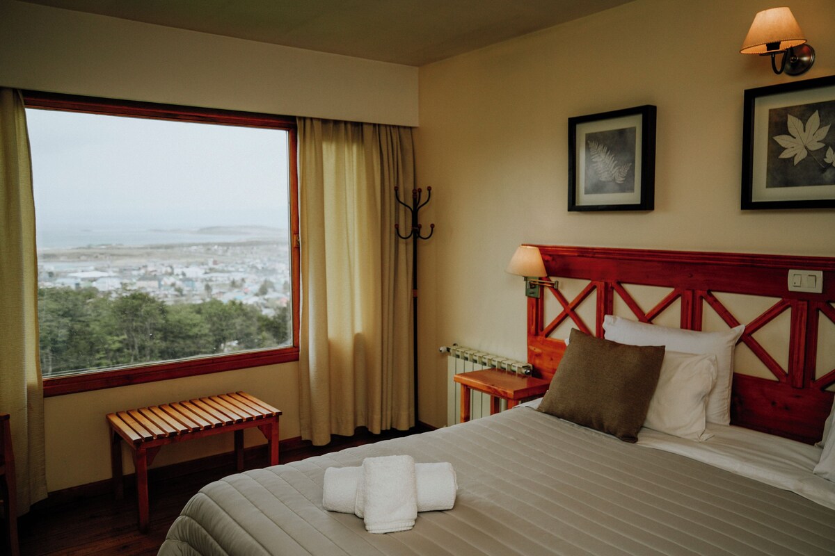 A hotel room with a large window, a bed with neatly folded towels, a wooden bench, and a view of a cityscape.