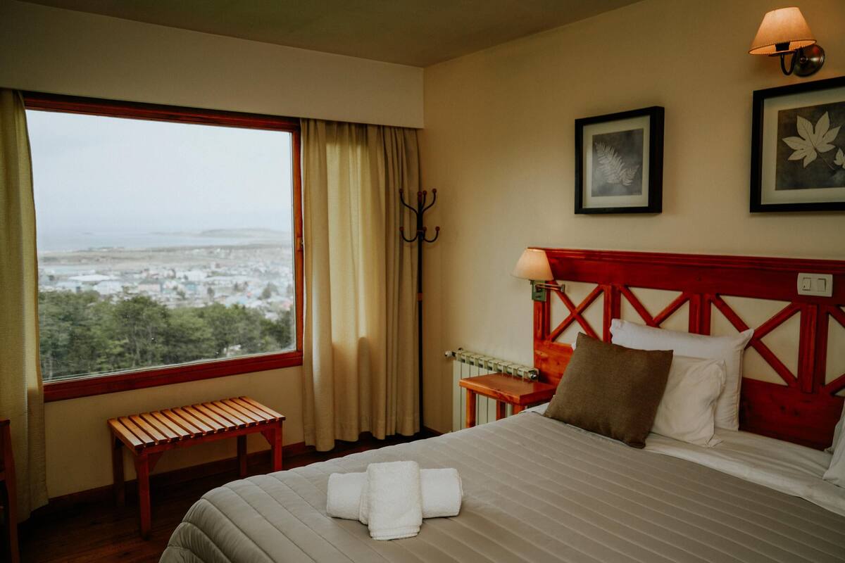 Une chambre d’hôtel avec un lit, une banquette, une vue sur la ville, un porte-manteau et une lampe.