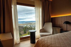 Una habitación de hotel con un ventanal grande, una cama, una silla, una mesita y una lámpara.
