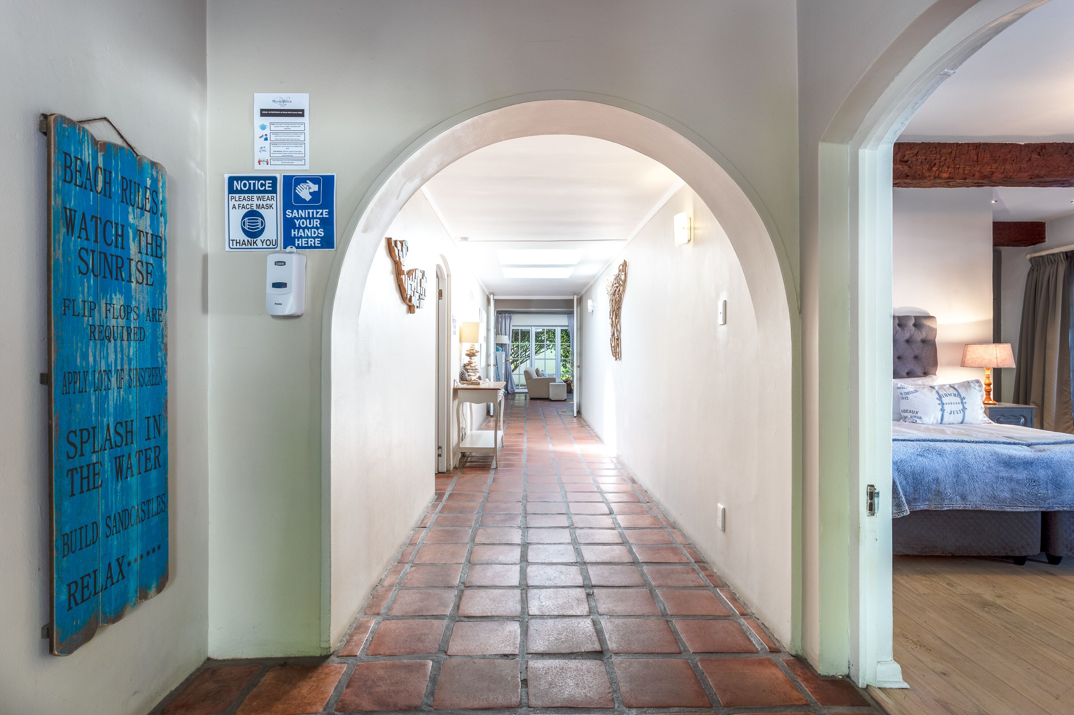 A hallway with a tiled floor leading to a bedroom with a bed and a nightstand. There are signs on the wall, including one that says "BEACH RULES WATCH THE SUNRISE" and another that says "SANITIZE YOUR HANDS HERE."