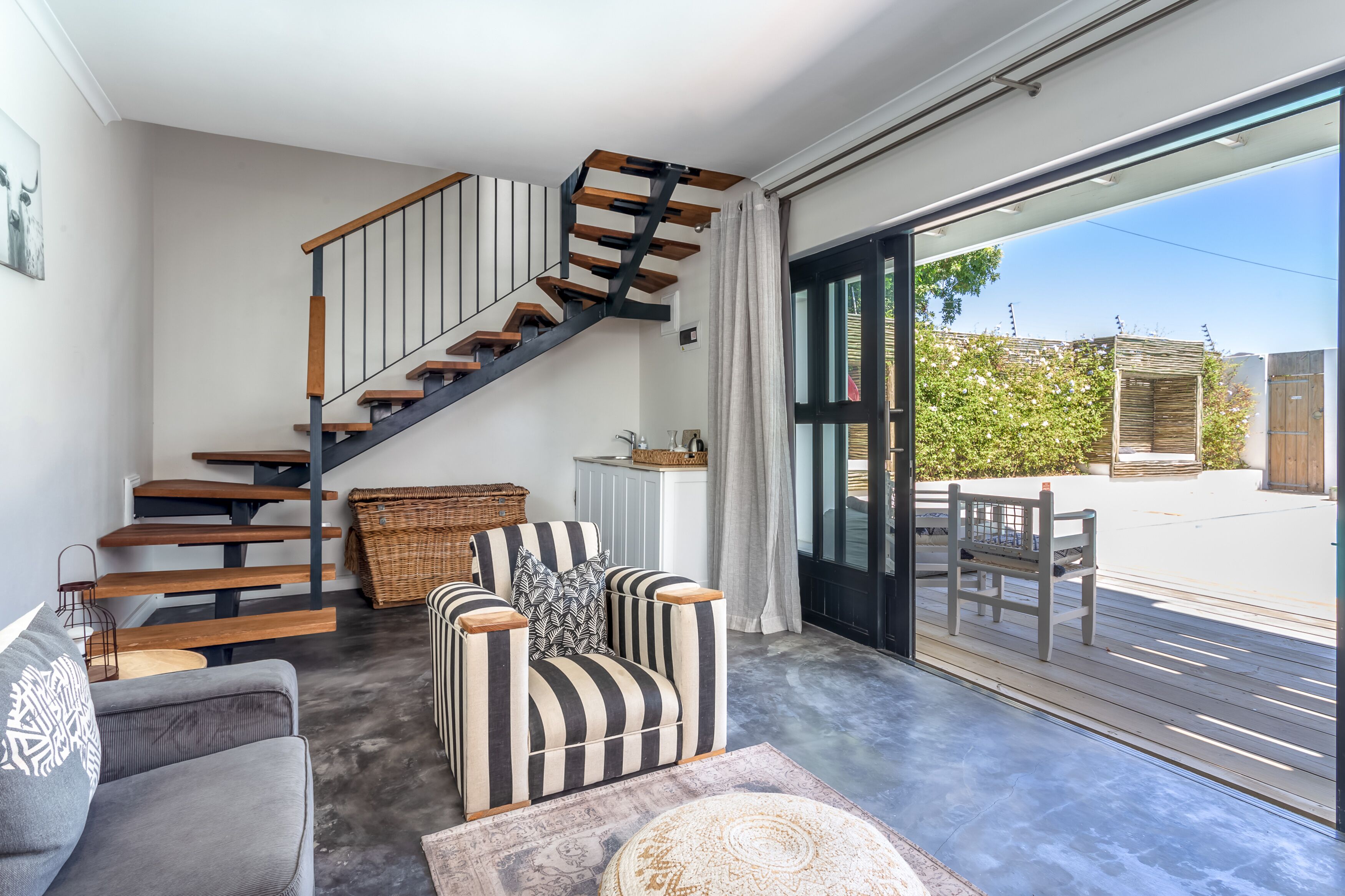 A modern interior with a wooden staircase, a grey sofa, striped armchairs, and a view of the outdoors through large windows.