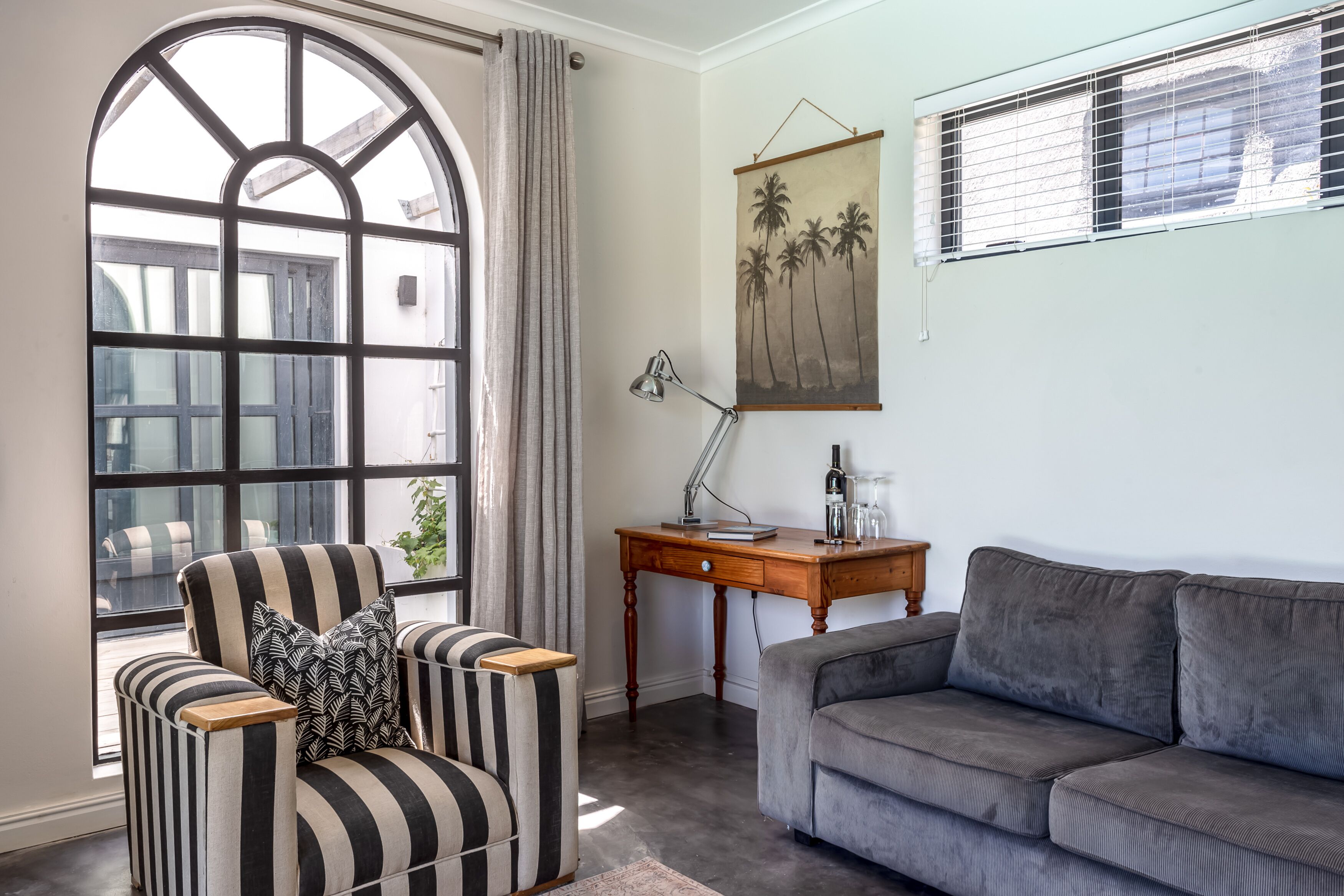 A living room with a large arched window, a striped armchair, a grey sofa, a wooden side table with a lamp, and a wall-mounted picture of palm trees.