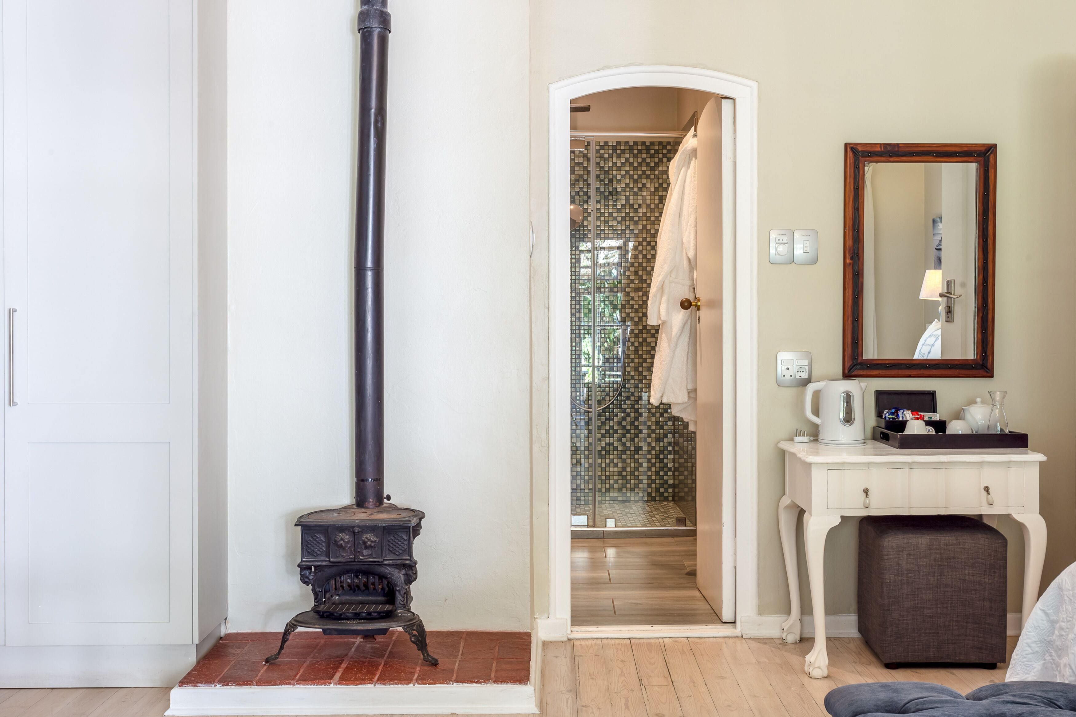A room with a white door, a black stove, a shower with mosaic tiles, a white vanity with a mirror, and a brown ottoman.