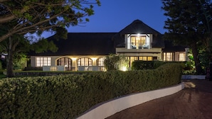 A large, modern house with a well-lit entrance, a curved driveway, and a neatly trimmed hedge. - Whale Rock Luxury Lodge (Hermanus)