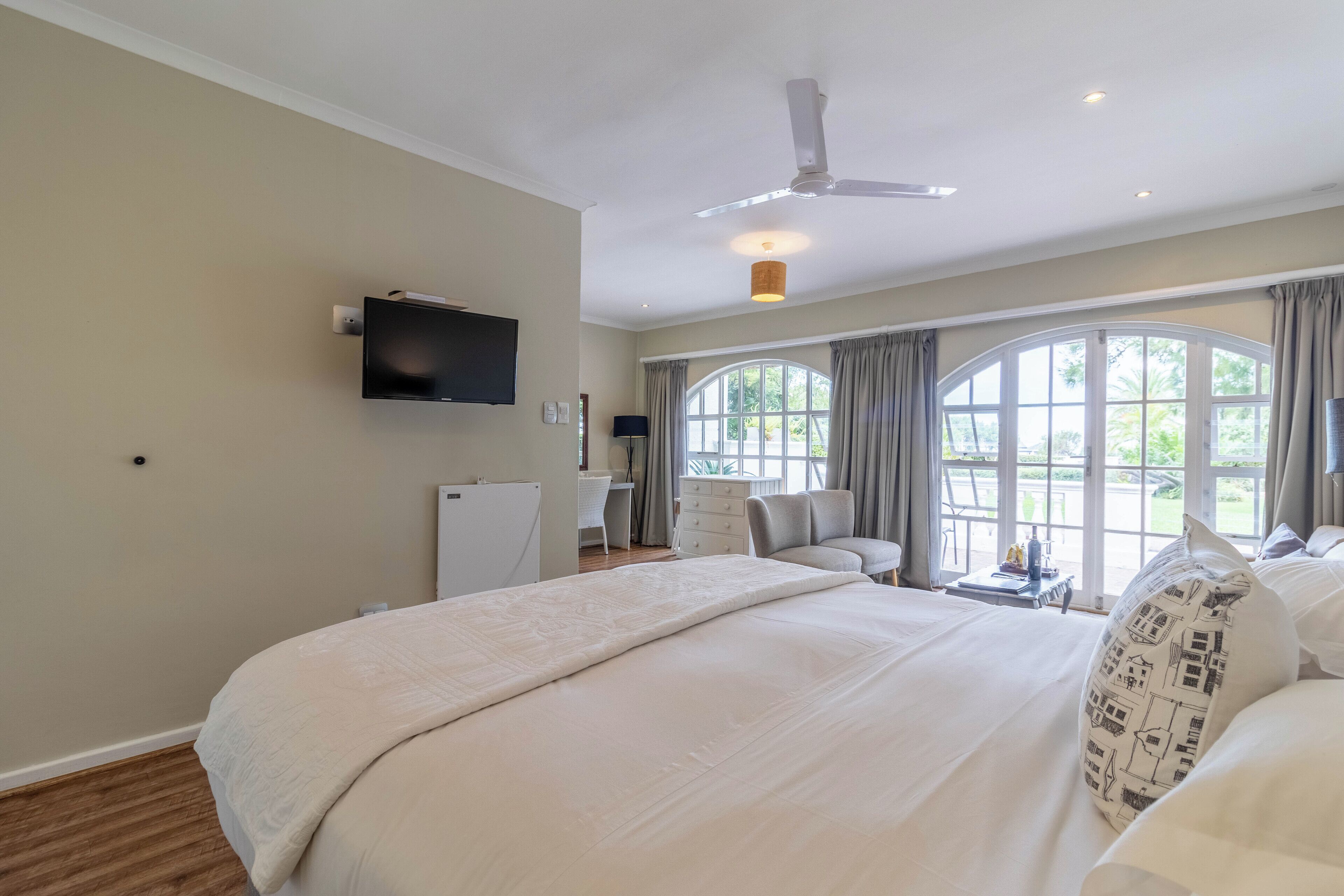 A spacious bedroom with a large bed, a ceiling fan, a flat-screen TV, and a view of the outdoors through large windows.