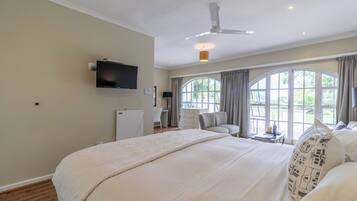 A spacious bedroom with a large bed, a ceiling fan, a flat-screen TV, and a view of the outdoors through large windows.