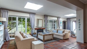 A spacious living room with two sofas, a coffee table, and a fireplace. Large windows with curtains provide natural light and views of greenery outside. - Whale Rock Luxury Lodge (Hermanus)