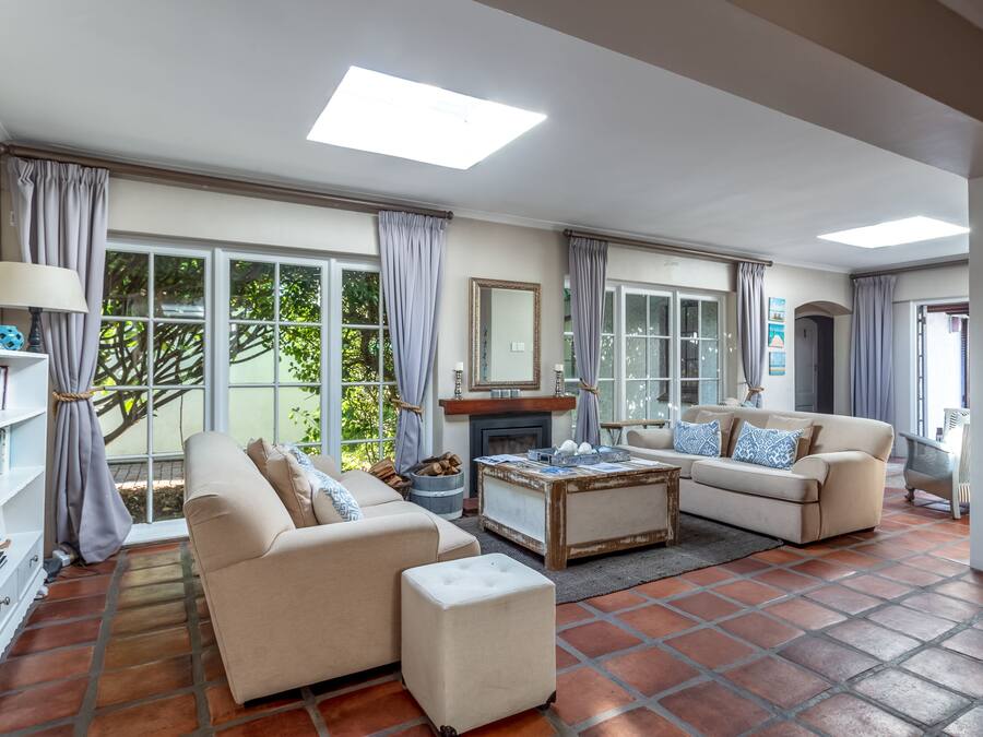 Un salon spacieux avec deux canapés, une table basse et une cheminée. De grandes fenêtres, ornées de rideaux, laissent entrer la lumière naturelle et offrent une vue sur la verdure environnante.