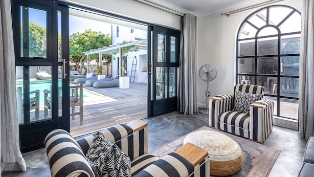 A spacious living area with a large sliding glass door leading to an outdoor deck, a cozy seating area with striped upholstered chairs, and a fan on the wall. - Whale Rock Luxury Lodge (Hermanus)