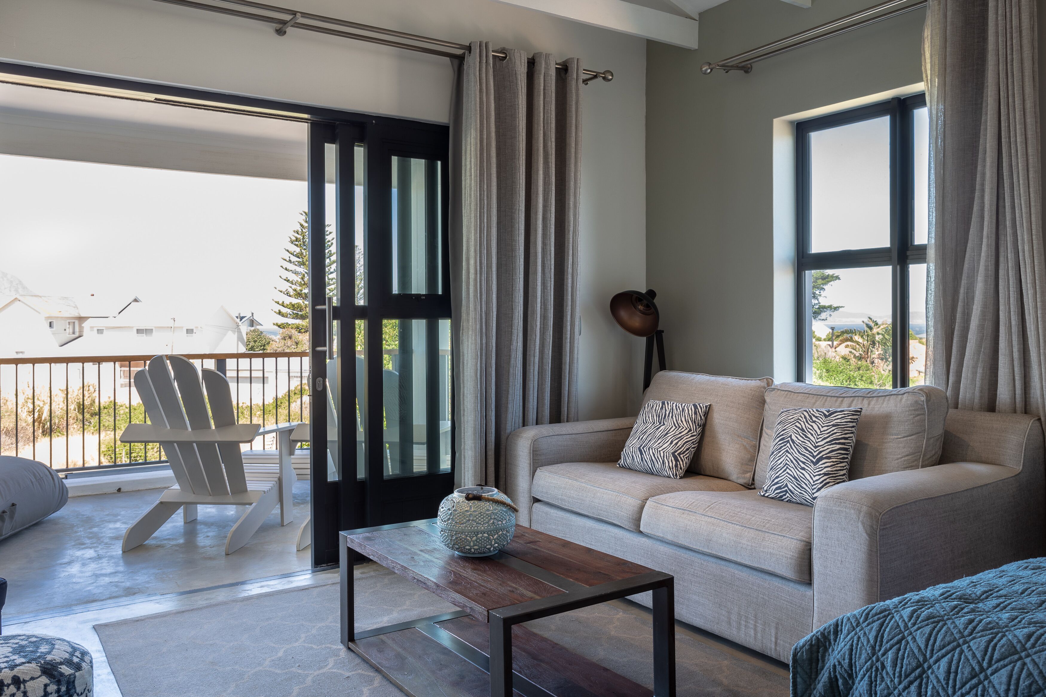 A modern living room with a sofa, a coffee table, and a chair. There is a large window with curtains, and a view of the outside.