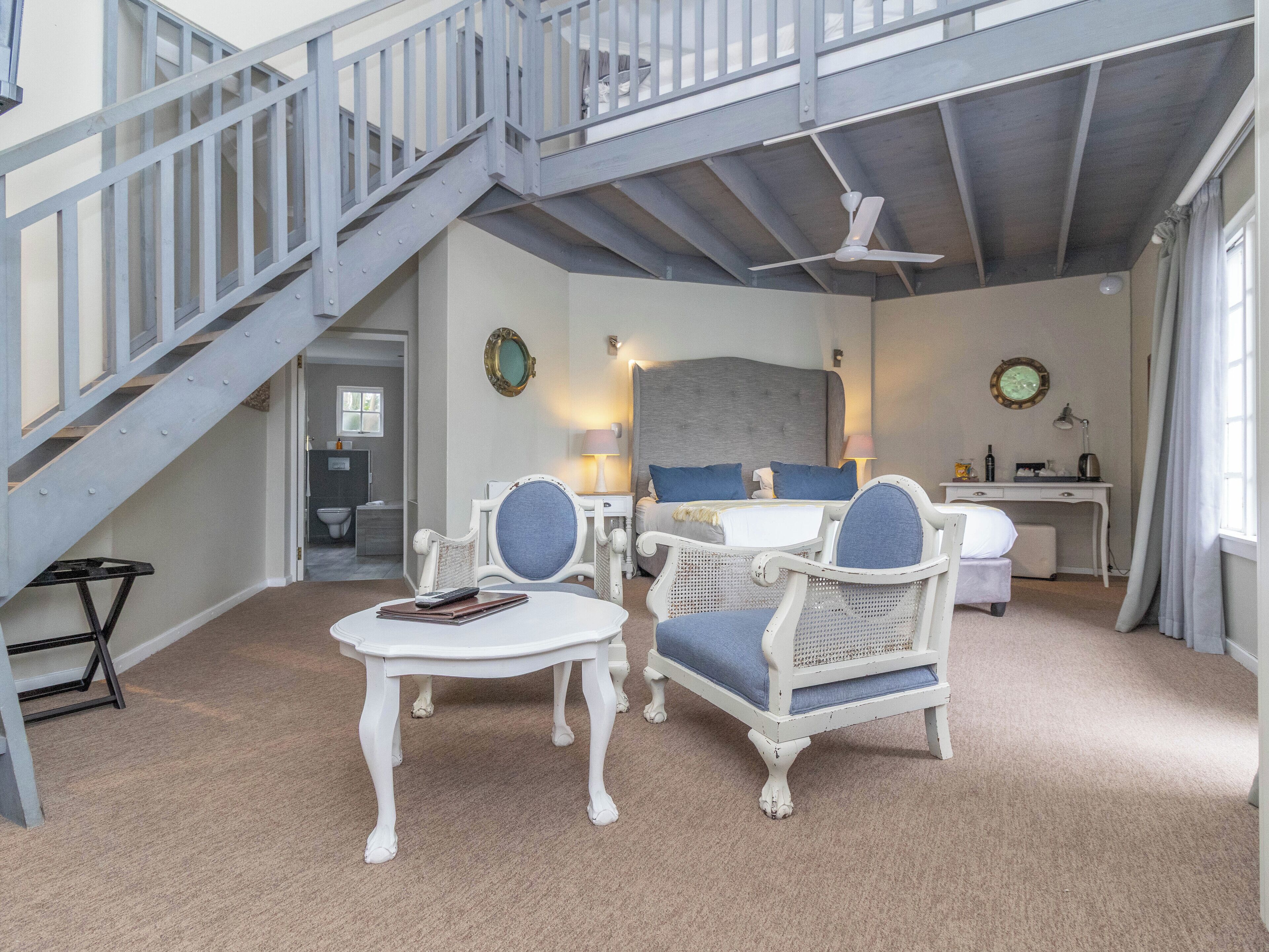 A room with a staircase, a bed with blue bedding, a desk, and two wicker chairs.
