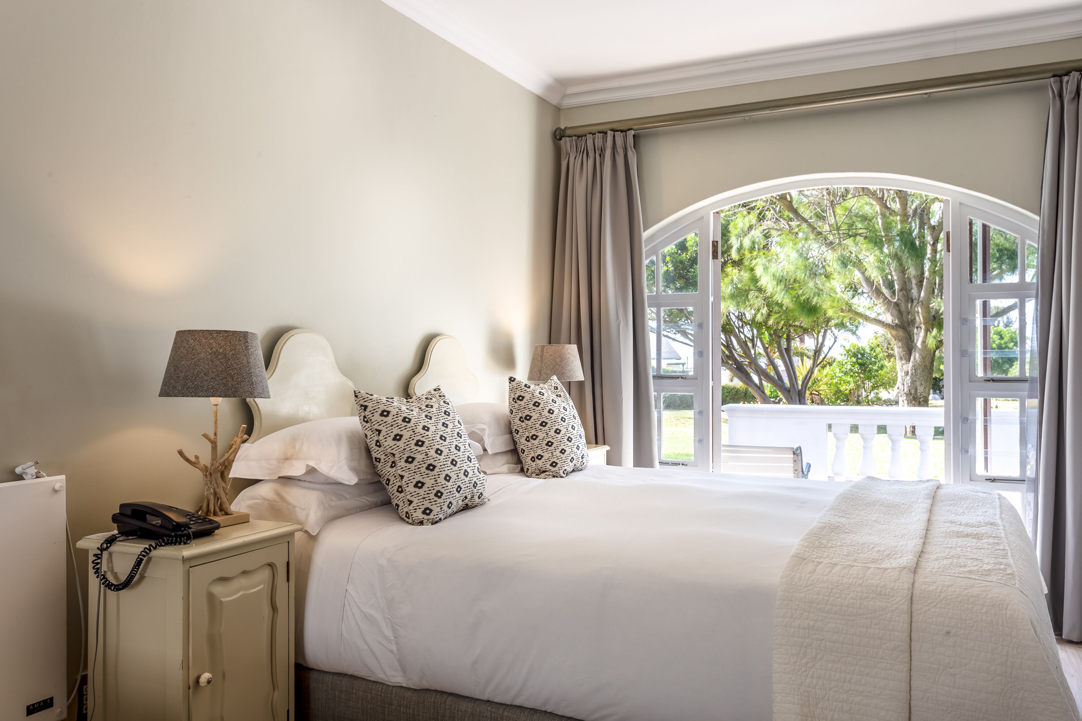 Habitación de hotel con una cama grande, una mesita de noche con lámpara, un teléfono y vistas a árboles a través de una ventana de arco.
