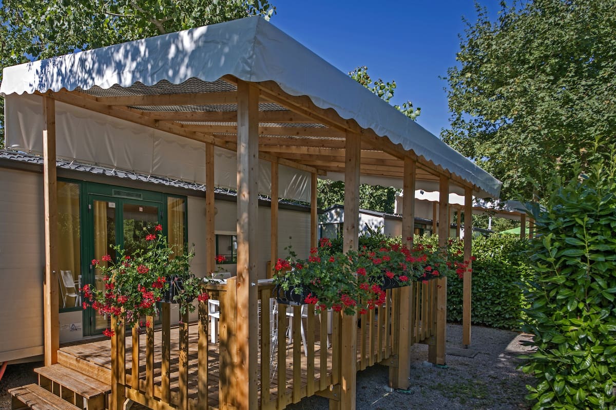 Une pergola en bois surmontée d’une toile blanche, une terrasse en bois et des fleurs rouges.