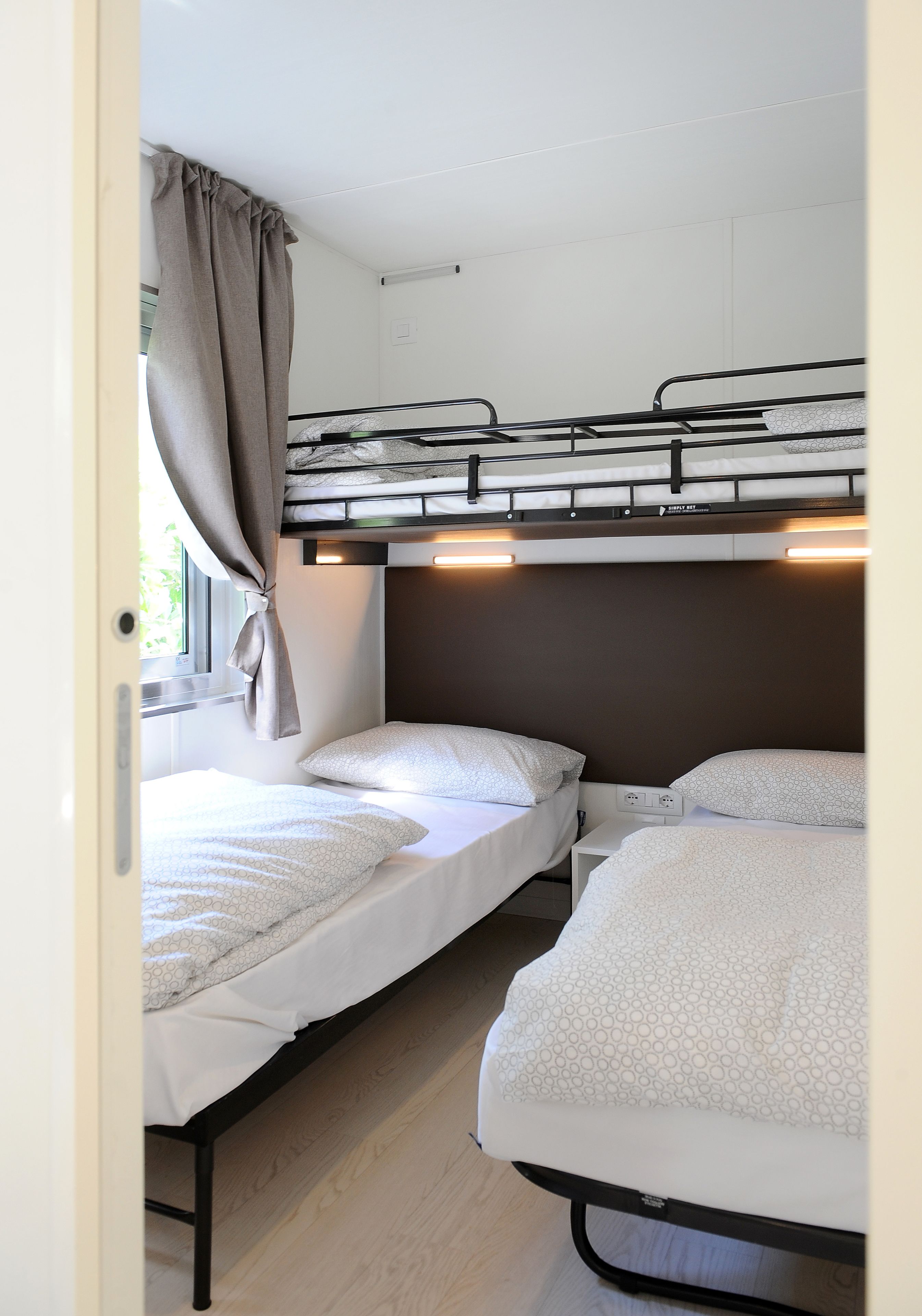 Une chambre compacte pour camping-car, dotée de deux lits superposés, d’une fenêtre avec rideaux et d’un éclairage intégré.