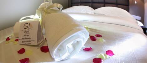 Un lit bien fait, avec un oreiller blanc et une serviette roulée, un petit panneau blanc indiquant les coordonnées de l’hôtel, et des pétales de rose éparpillés.