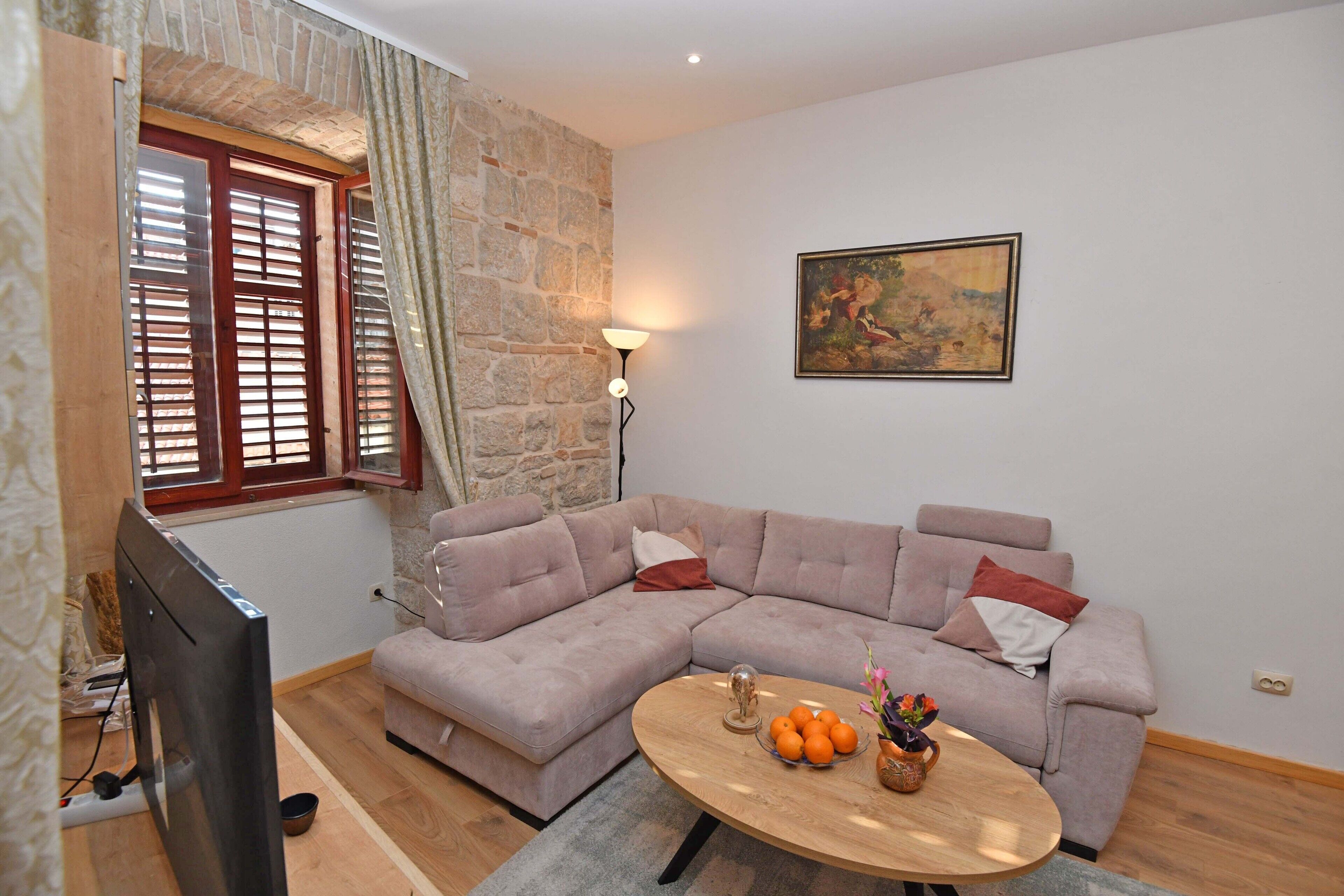 A living room with a sectional sofa, a round coffee table with oranges and flowers, a painting on the wall, and a stone wall.