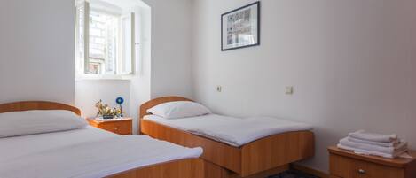 Un dormitorio con dos camas de madera, una mesita de noche pequeña con una lámpara y una ventana con un persiano abierto.