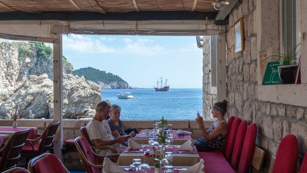 Un restaurant en bord de mer, avec des murs en pierre, des sièges rembourrés de couleur rouge et une vue sur la mer et un navire.