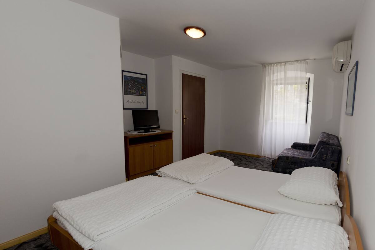 A hotel room with a large bed, a desk with a computer, a door, and a window with sheer curtains.