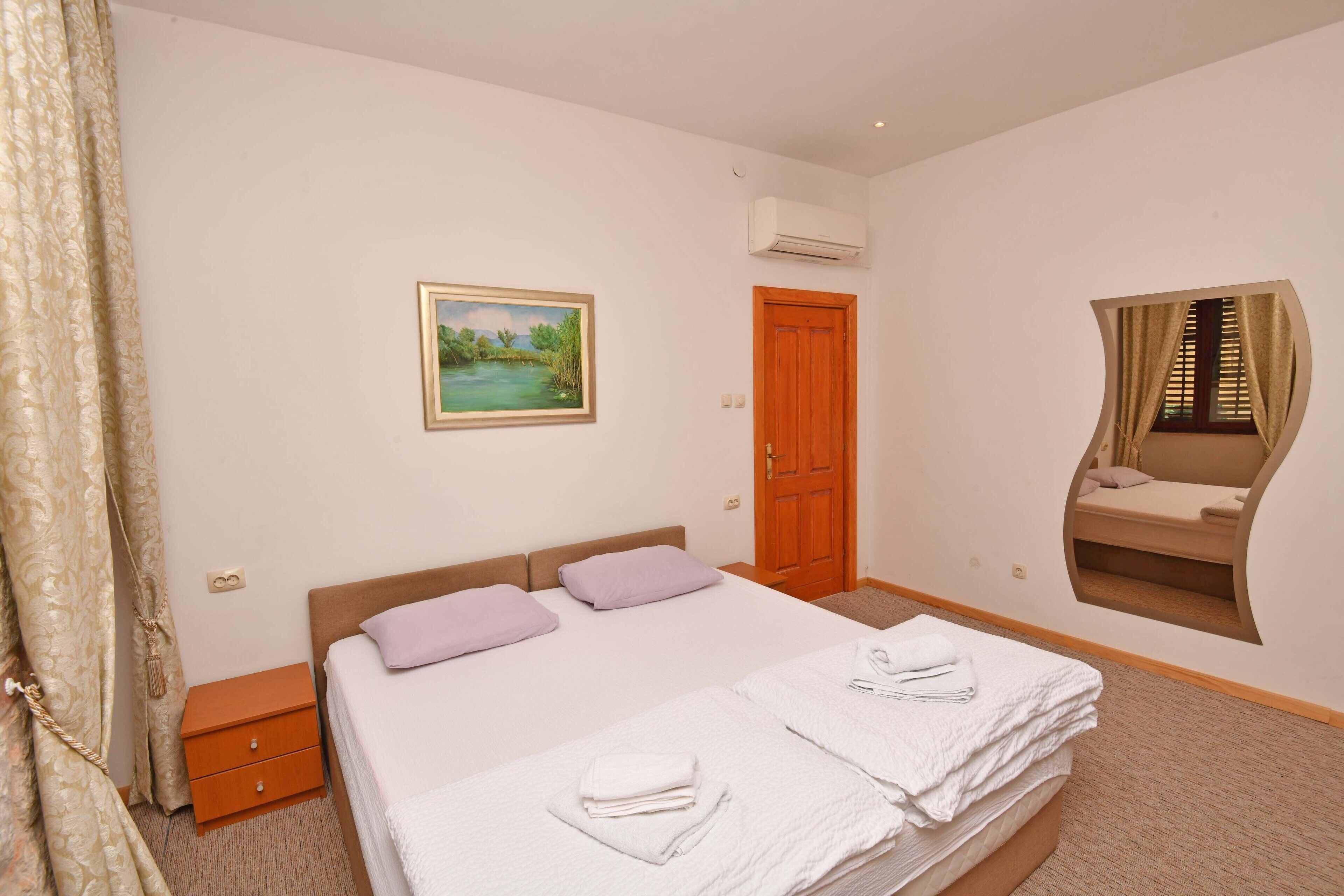 A bedroom with a bed, a nightstand, a wooden door, an air conditioning unit, a painting, and a built-in window seat with curtains.