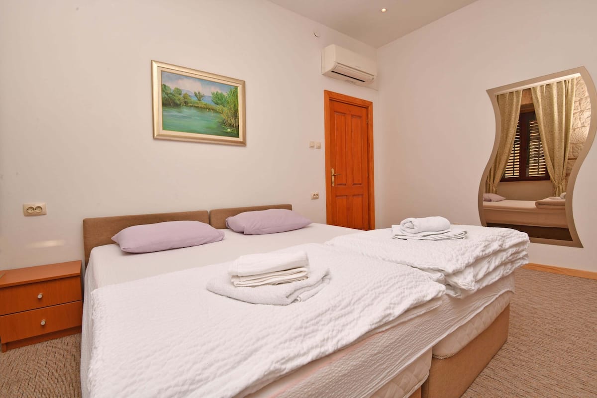 A bedroom with a bed, a nightstand, a painting, an air conditioning unit, and a window with curtains.
