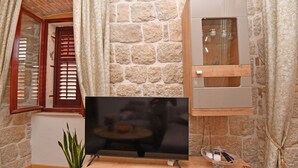 Une pièce avec un mur en pierre, un meuble en bois doté de portes en verre, un téléviseur à écran plat, une petite table avec une plante et une fenêtre avec des volets en bois.