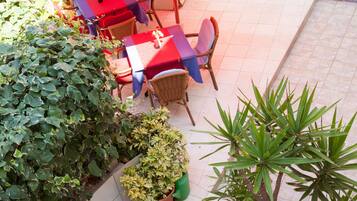 Une terrasse extérieure avec des tables et des chaises, entourée de plantes en pot et d’un grand parasol.
