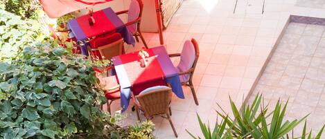 Patio esterno con tavoli e sedie, circondato da piante in vaso e un grande ombrellone.