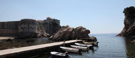 Cena costeira com um cais de pedra, várias embarcações pequenas ancoradas e uma grande formação rochosa.